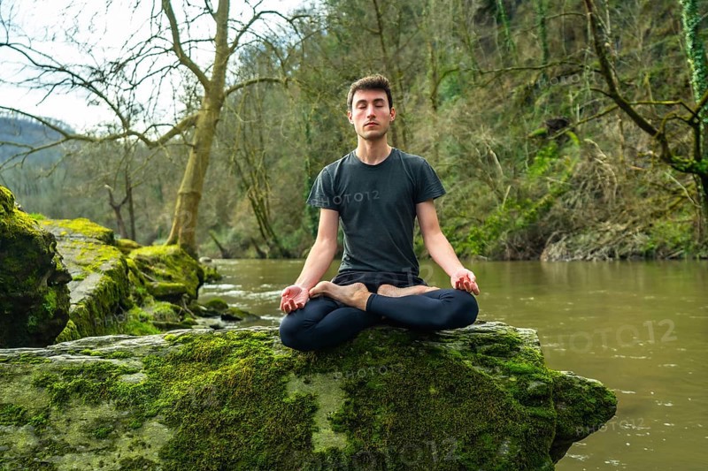 Yoga instructor meditating in lotus position on a mossy rock by a tranquil river, surrounded by lush nature, promoting mindfulness and serenity