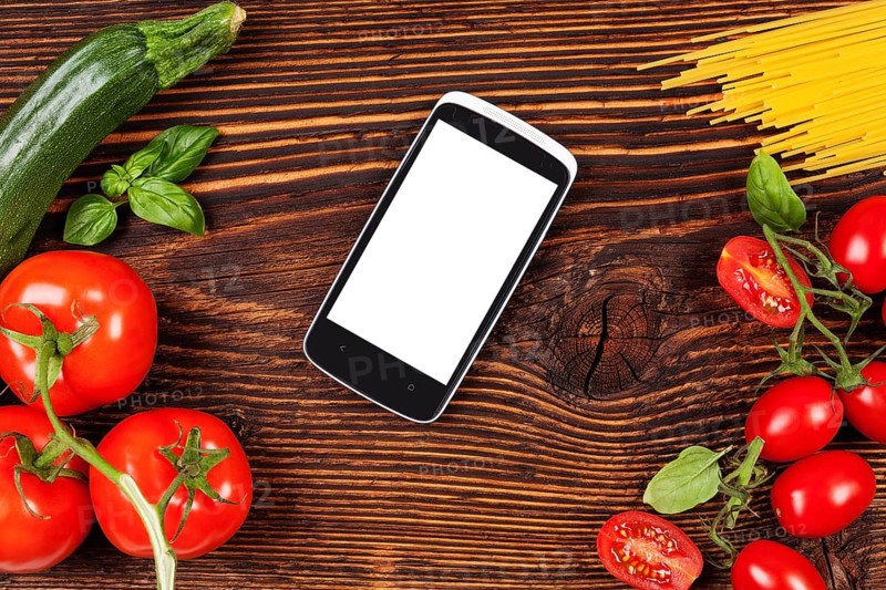 Fresh vegetables and pasta on wooden table with smartphone and recipe on screen. Groceries, recipe and cooking. Modern food preparation