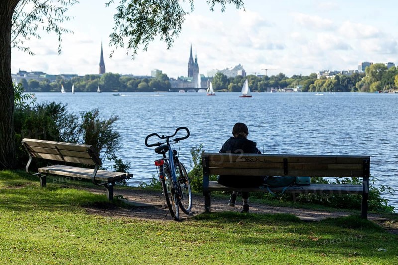 Leisure activities in summer, bicycle and woman on park bench, behind them sailing boats in front of blue sky on the Alster, Hamburg, Germany