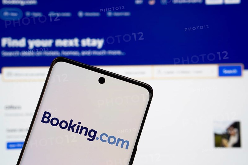 Booking.com logo is displayed on a smartphone screen, with the booking.com website visible on a computer screen in the background