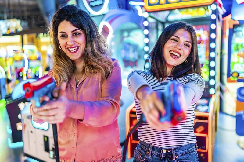 Two young women enjoying their free time playing together in an amusement arcade, holding toy guns and having fun