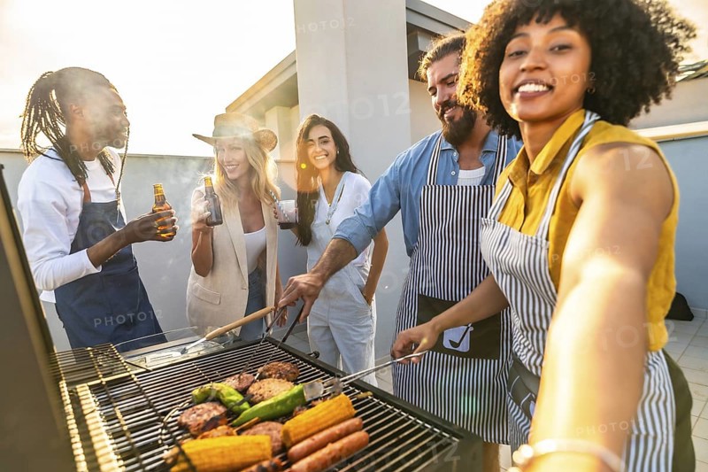 Cheerful multi ethnic friends are having fun, drinking beer and taking selfie while preparing barbecue on a rooftop at sunset