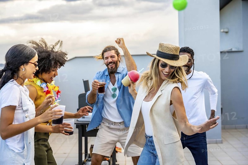 Cheerful multi ethnic friends dancing joyfully and sipping cocktails while celebrating together at an exciting rooftop party during sunset