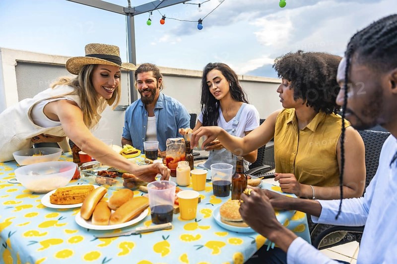 Happy multi ethnic friends having lunch together on urban rooftop, eating hot dogs, burgers, and drinking beer