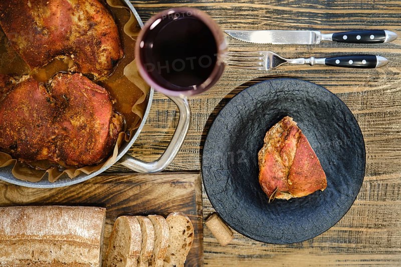 Overhead view of roasted meatin a plate and baking pan served with freshly sliced bread and a glass of red wine