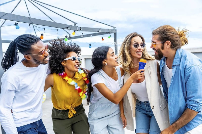 Five cheerful friends singing karaoke, holding a microphone and enjoying a rooftop party during summertime