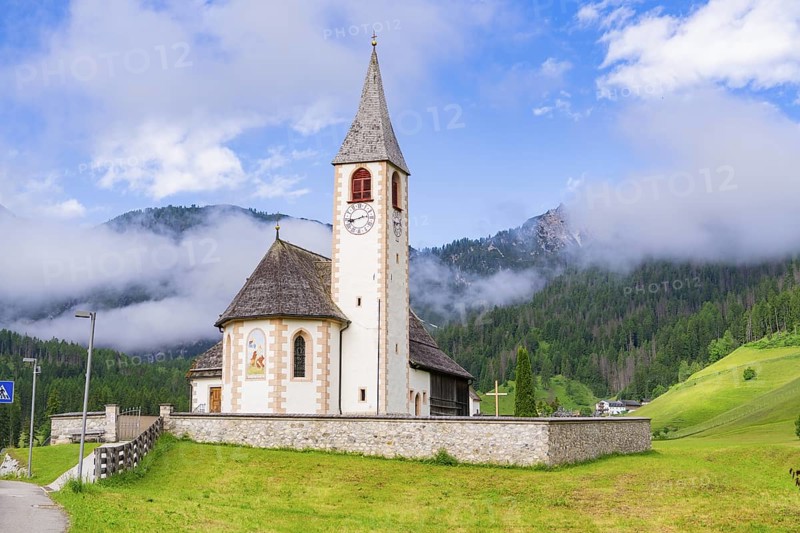 Church with tower and clock, surrounded by meadows and mountains in an alpine landscape, Dolomites, South Tyrol, Italy
