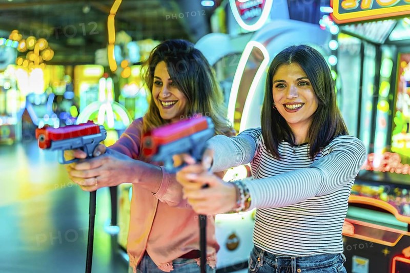 Two happy women enjoying their free time playing together in an amusement arcade holding toy guns