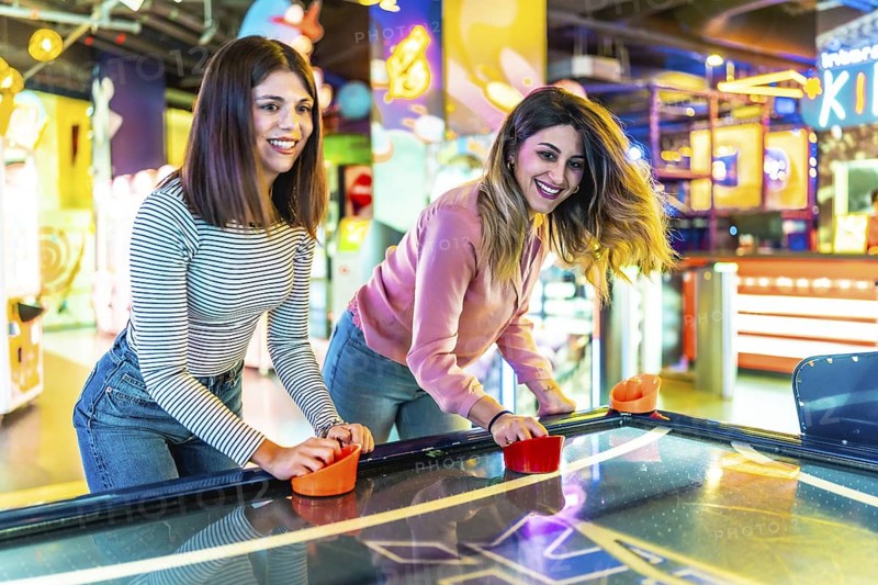 Two female friends enjoying their free time playing air hockey in a colorful and modern amusement arcade