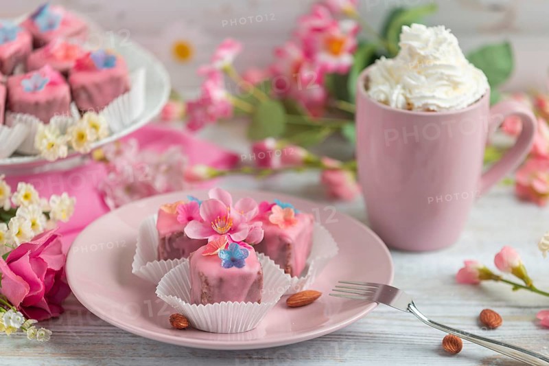 Pink petit fours with flower decoration on a plate next to a cup of coffee with cream