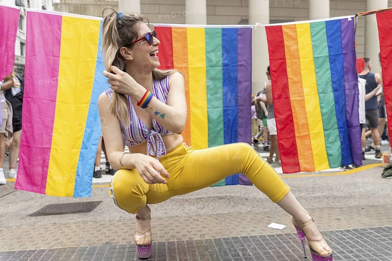 Young woman wearing colorful clothes and rainbow bracelet crouching in front of rainbow and pansexual flags, touching her hair and laughing during gay pride parade