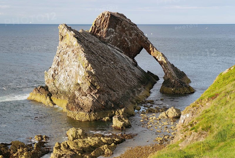 Large rock formation in a calm sea illuminated by the setting sun, Remarkable rock formation jutting into the sea under a blue sky with few clouds, Bow Fiddle Rock, Aberdeenshire, Buckie, Scotland, United Kingdom