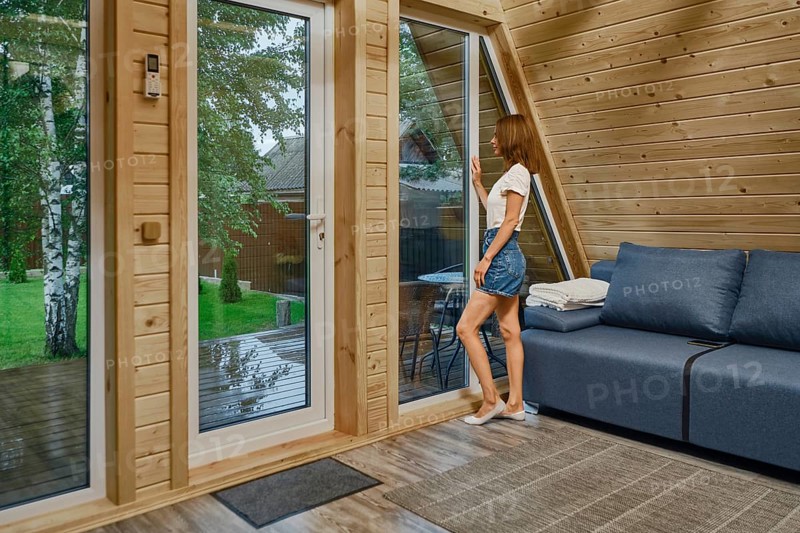 Cute woman stands by large glass window of a wooden cabin, looking out at the lush greenery after the rain