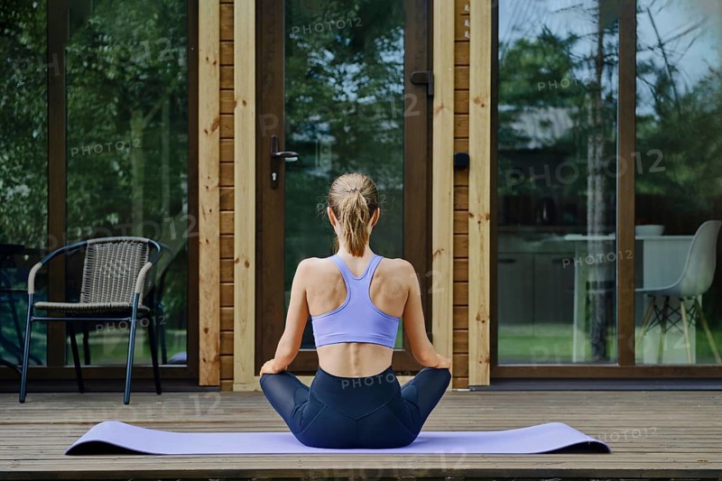 Back view of a woman sits in lotus position on a yoga mat placed on a wooden terrace of tiny house