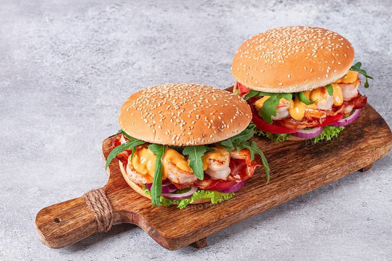 Two shrimp burgers and buns with sesame seeds, bacon, lettuce, tomatoes and onions, On a wooden board