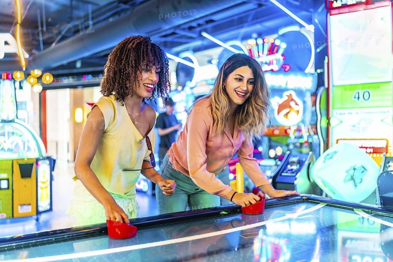 Two young women enjoying a friendly game of air hockey in a vibrant arcade, illuminated by colorful neon lights