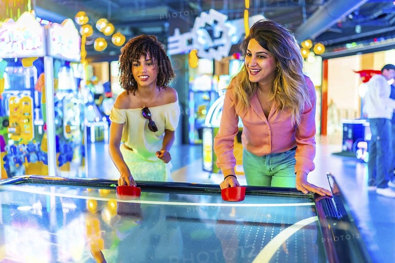 Two female friends enjoying a lively game of air hockey in a vibrant amusement arcade filled with colorful lights and excitement