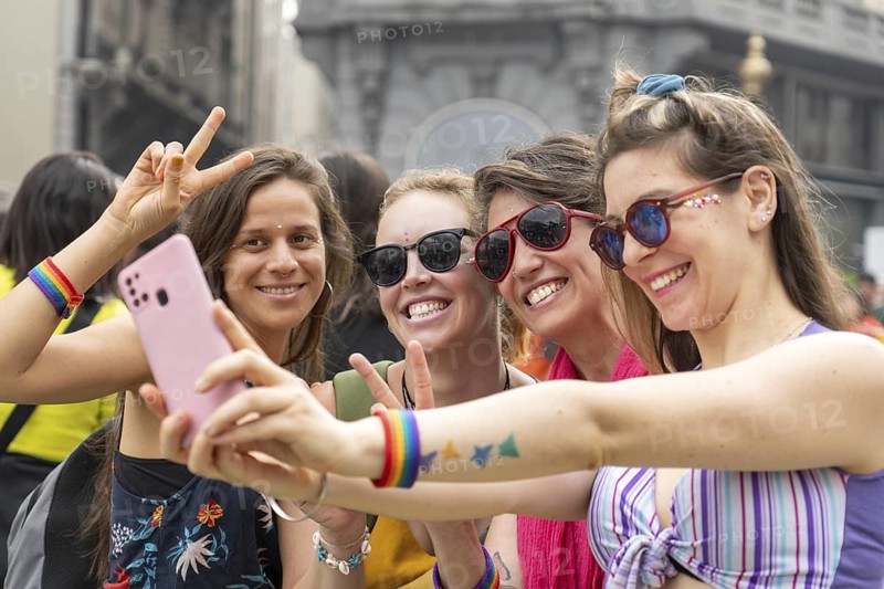 Four joyful women wearing colorful rainbow bracelets are capturing a selfie with a smartphone during a vibrant pride parade, flashing peace signs and beaming with happiness