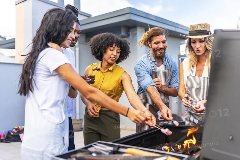 Cheerful friends roasting marshmallows on skewers over a barbecue grill during a rooftop party
