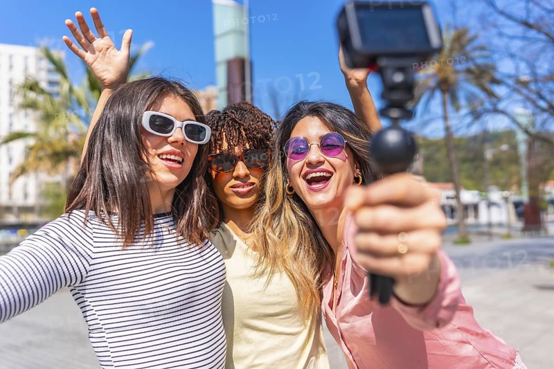 Three cheerful women holding action camera and waving at camera while vlogging their summer vacation in a sunny city