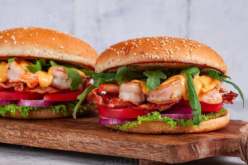 Two shrimp burgers and buns with sesame seeds, bacon, lettuce, tomatoes and onions, On a wooden board