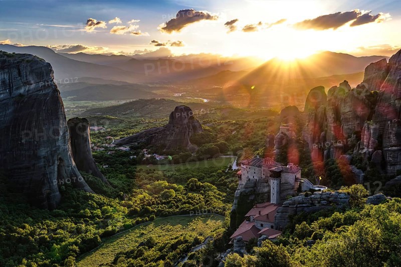 Iconic view of Meteora valley at sunset. Sun setting behind distant mountains, sun rays fall on valley, cliffs, rocks, clifftop Moni Agias Varvaras Roussanou nunnery, blue sky and beautiful clouds. Natural lens flares. Greece, UNESCO World Heritage