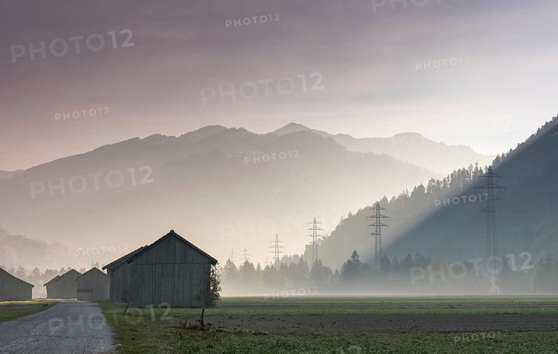 View of the Praettigau Valley near Klosters in Switzerland on an early morning with farm fields and wooden barns and power lines and mountains in silhouette behind in late autumn