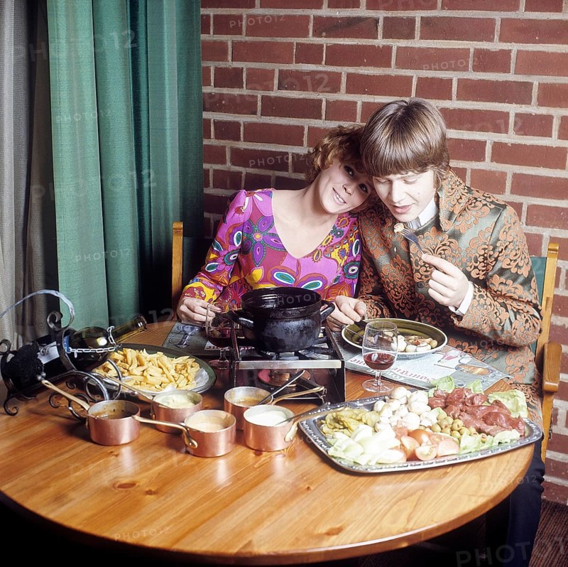 Fondue. A dish that had its breakthrough in the 60s and 70s. A fondue pot with hot oil stands in the middle of the table and you dip meat into it to cook and eat yourself. She is wearing a very colorful and patterned long sleeved blouse that was typical of the late 60's and 70's. Him in a pop inspired jacket not unlike the clothing that singers wore at the time. Sweden 1970. Conard ref CV49-5