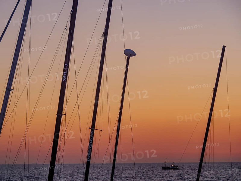 Masts in the foreground and a small boat in the sea at sunset, the sky is bathed in orange tones, De Haan, Flanders, Belgium