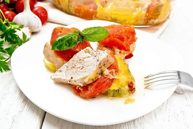 Vegetable casserole with chicken breast, red tomatoes, zucchini and spices in a plate and glass roasting pan on a towel, parsley, garlic and hot peppers on background of light wooden board