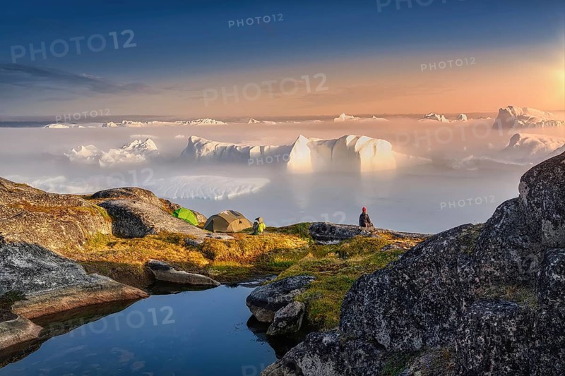 Large glaciers in the bay by the rocky shore in the light of the setting sun with fog over the water, woman in a red cap