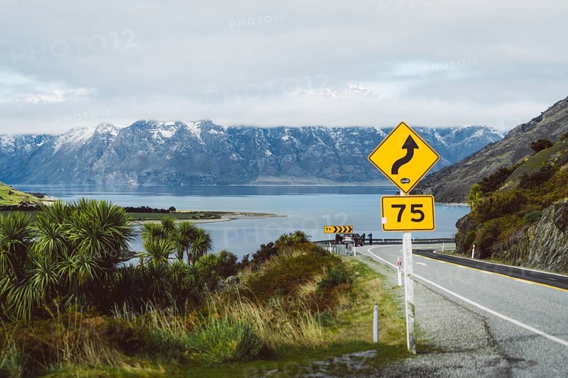 Road with danger signs leads along the lake in front of snow-covered mountains, Wanaka, New Zealand
