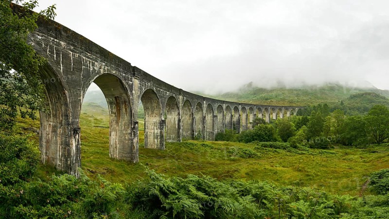 High resolution panorama of Glenfinnan railway viaduct (location from Harry Potter movie) shot from side, on overcast day with grey sky and lot of green grass and trees around. Inverness, Scotland, United Kingdom