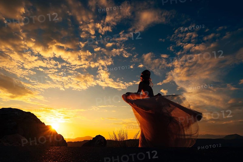 Bride in a fluttering dress stands on a mountain against the backdrop of a sunset sky.