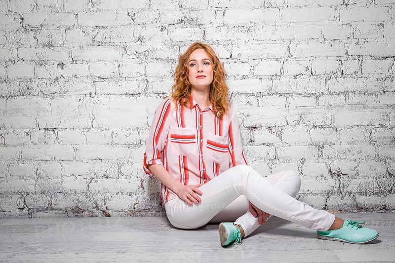 Young beautiful caucasian woman student is sitting on the floor near a white brick wall. Girl with red long curly hair with freckles on her face in bright summer clothes