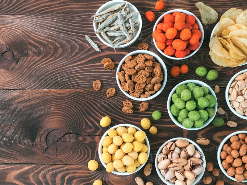 Assortment of different snack for beer, wine, party. Peanuts in coconut glaze, green vasabi, red spicy chilli, yellow cheese glaze, chips, pistachio, crackers, fish on brown wooden table. Copy space