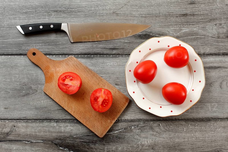 Table top view on three tomatoes on white porcelain plate with red dots, cutting board with tomato cut to half next to it, chef knife also laying on the gray wood desk