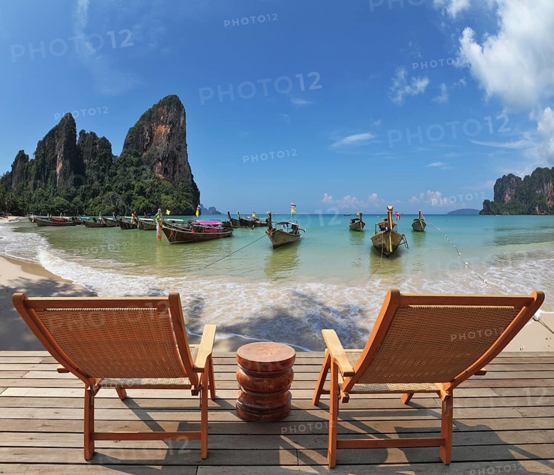 Cozy beach rest. Two wooden chaise lounges and small bedside table on a wooden scaffold. Native boats for sea walks expect tourists near the coast