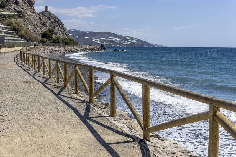 Promenade with wooden railings by the sea in front of the village with blue sky and sea, Castillo de Baños de Abajo, Polopos, Granada, Andalusia, Spain