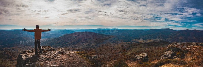 Panoramic view of mountain valley. Young man standing with hands widespread trying to embrace whole world. Man is a small part of astonishind mountain landscape with cloudy sky