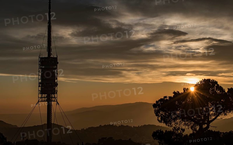 A tall communication tower stands silhouetted against a dramatic sunset with mountains and a tree in the foreground, Collserola tower, Barcelona, Catalonia, Spain