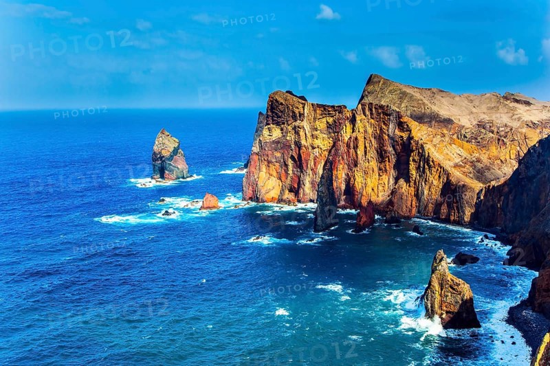 Island in the Atlantic. The coast of the island of Madeira - red rocks, azure surf. The blinding midday sun illuminates the rocky shore. The concept of active, ecological and photo tourism