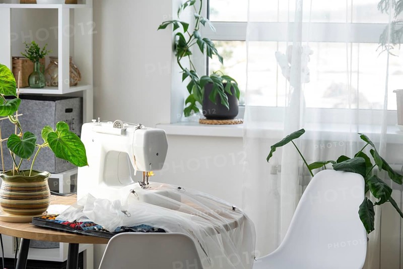 Sewing machine on a round table in the white interior of the house near the window with a transparent curtain, and a home plant in a pot, a modern interior. hobby, home business