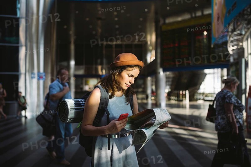 Theme travel and transportation. Beautiful young caucasian woman in dress and backpack standing inside train station terminal looking at electronic scoreboard holding phone, map paper hand navigation