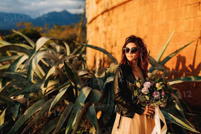 Bride in a beautiful pastel dress and sunglasses holds a bouquet of flowers in her hands against the background of agave bushes and the castle wall.