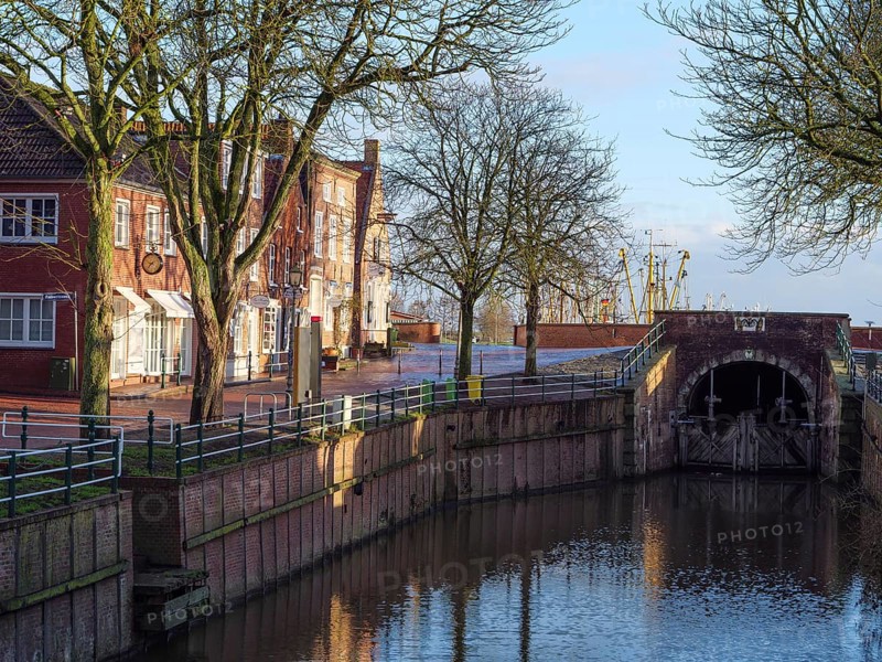 Urban scene with canal lined with trees and buildings. Bridge and reflection visible in the water, Greetsiel, East Frisia, Germany