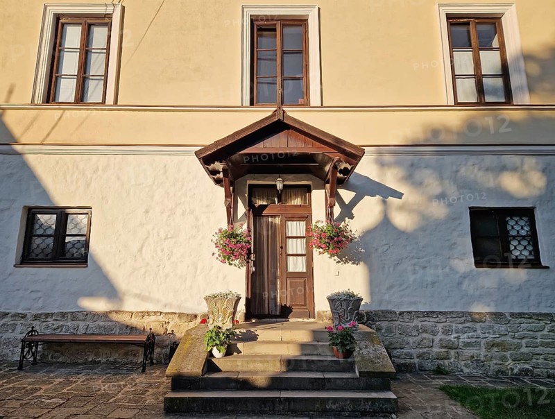 Rustic old building facade with porch and a charming wooden door, framed by two side windows and a small overhang. Stone steps lead up to the entrance, adorned with flower pots in bloom
