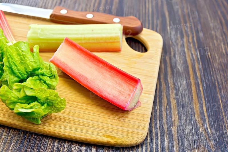 Rhubarb stalks with green leaves and a knife on a small planch on a wooden boards background