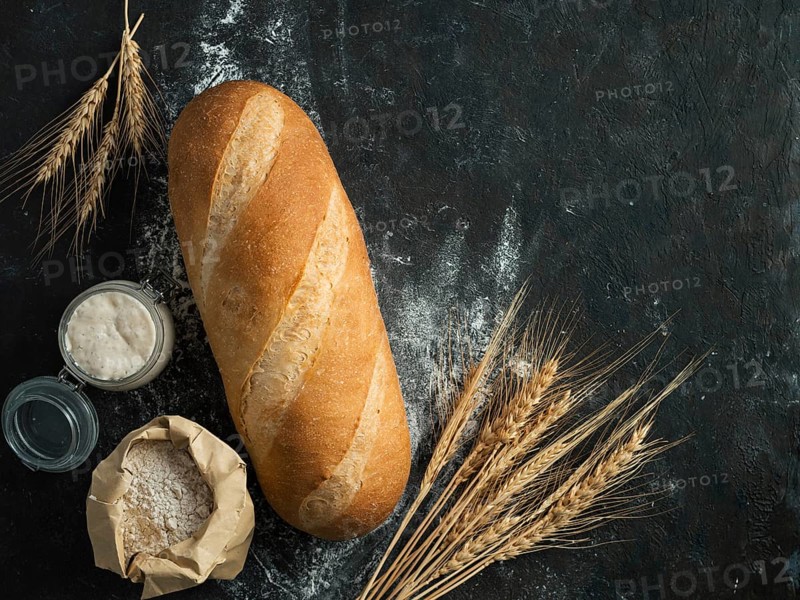 British White Bloomer or European sourdough Baton loaf bread on black background. Fresh loaf bread and glass jar with sourdough starter, floer in paper bag and ears. Top view. Copy space