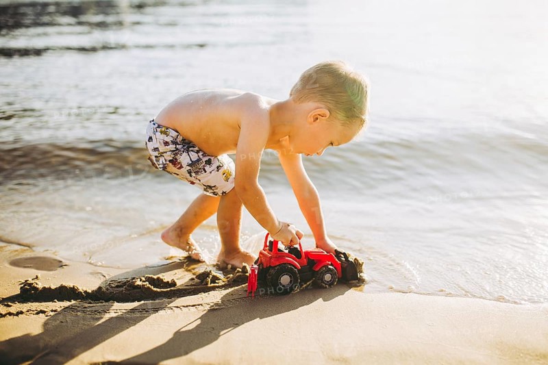Caucasian child boy plays toy red tractor, excavator on sandy beach by the river in shorts at sunset day